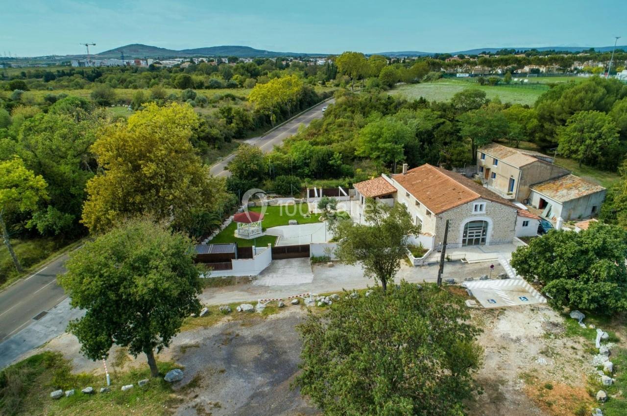 Vue aérienne d'une maison en pierre avec toit en tuiles, entourée de verdure et située près d'une route de campagne.