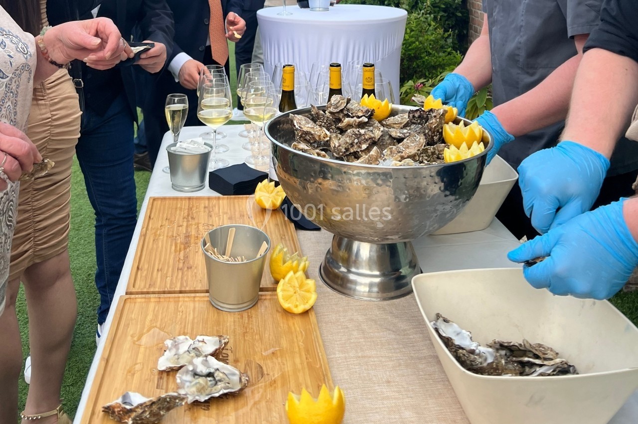 Des huîtres sont ouvertes et servies sur une table lors d'un événement en plein air avec des verres de vin en arrière-plan.