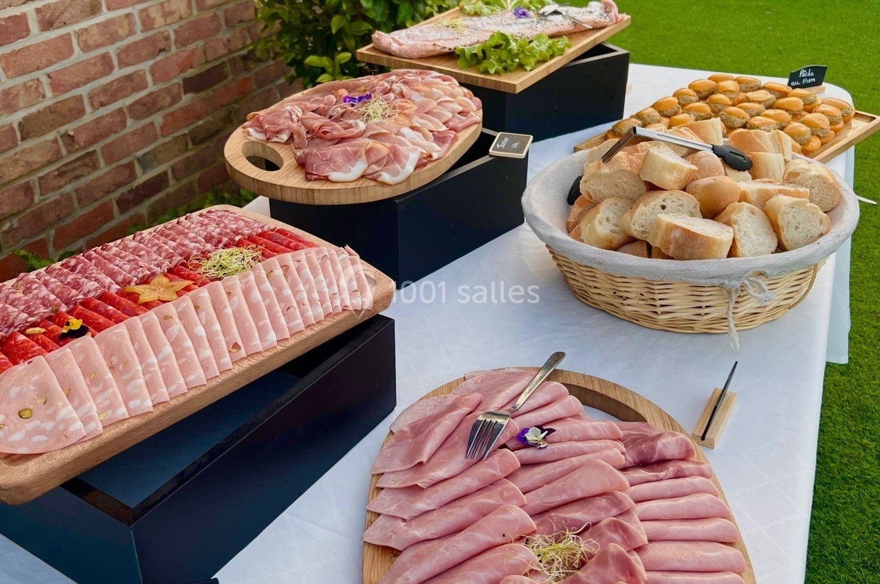 Plateau de charcuterie variée, pain et petits pains disposés sur une table en extérieur avec nappe blanche.