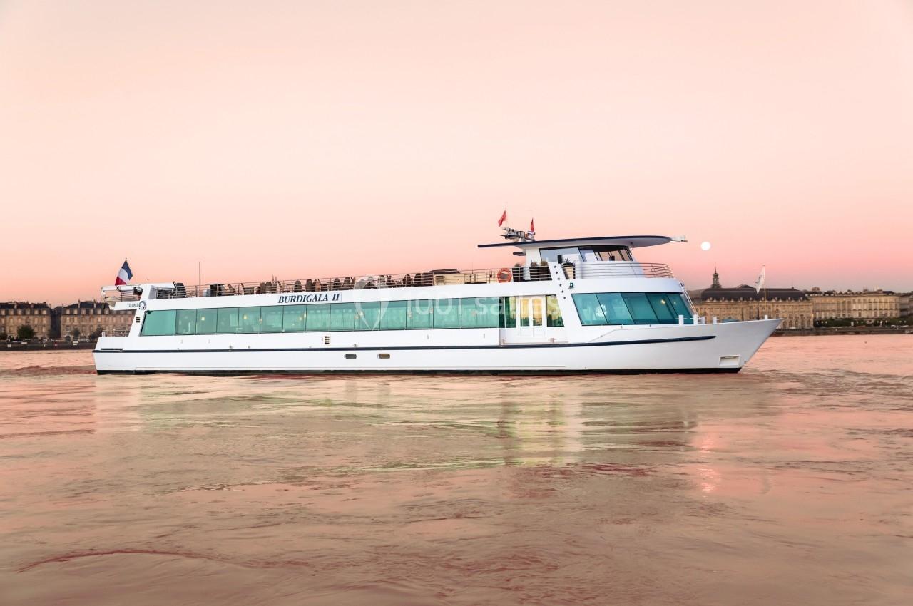 Bateau de croisière blanc naviguant sur une rivière au coucher du soleil, avec des bâtiments en arrière-plan.