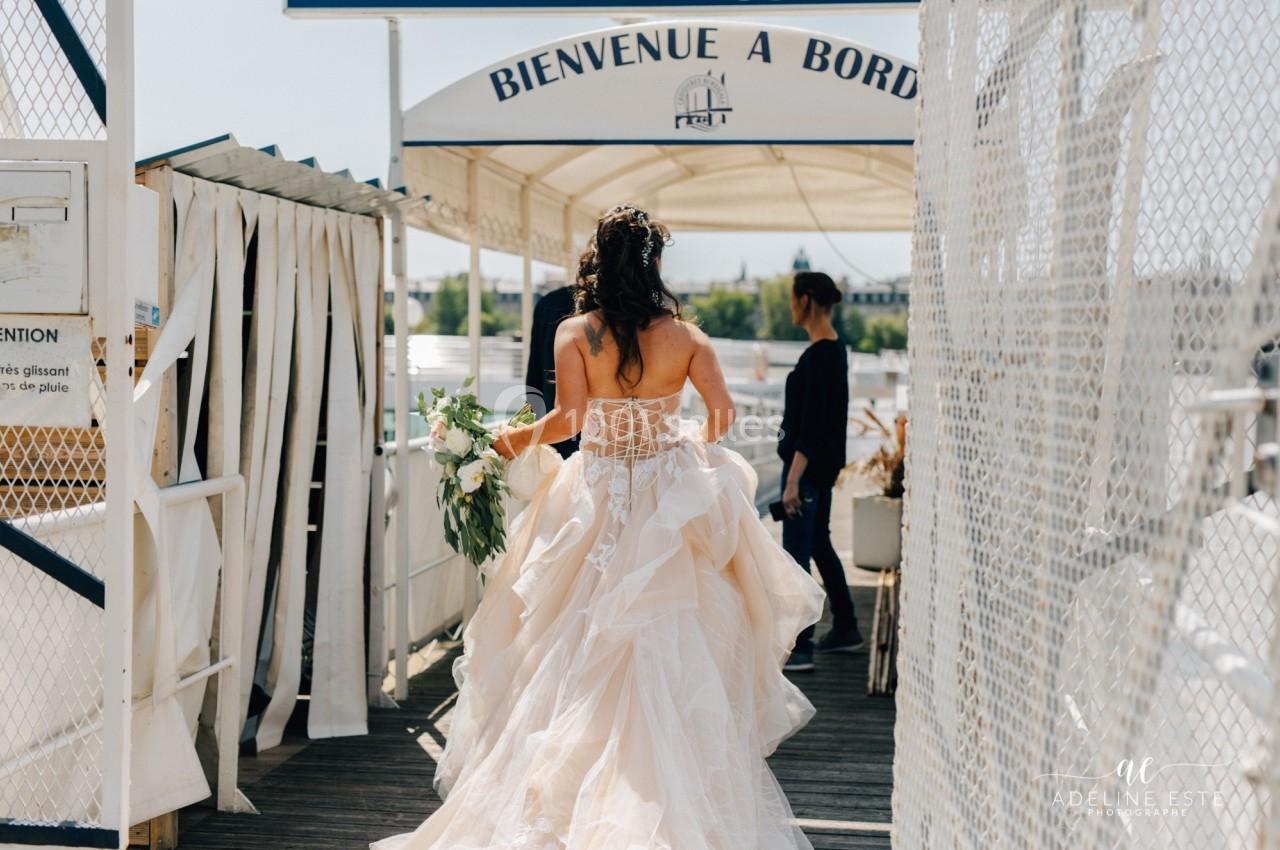 Une femme en robe de mariée marche sur un ponton, tenant un bouquet, sous une arche marquée ’Bienvenue à bord’.