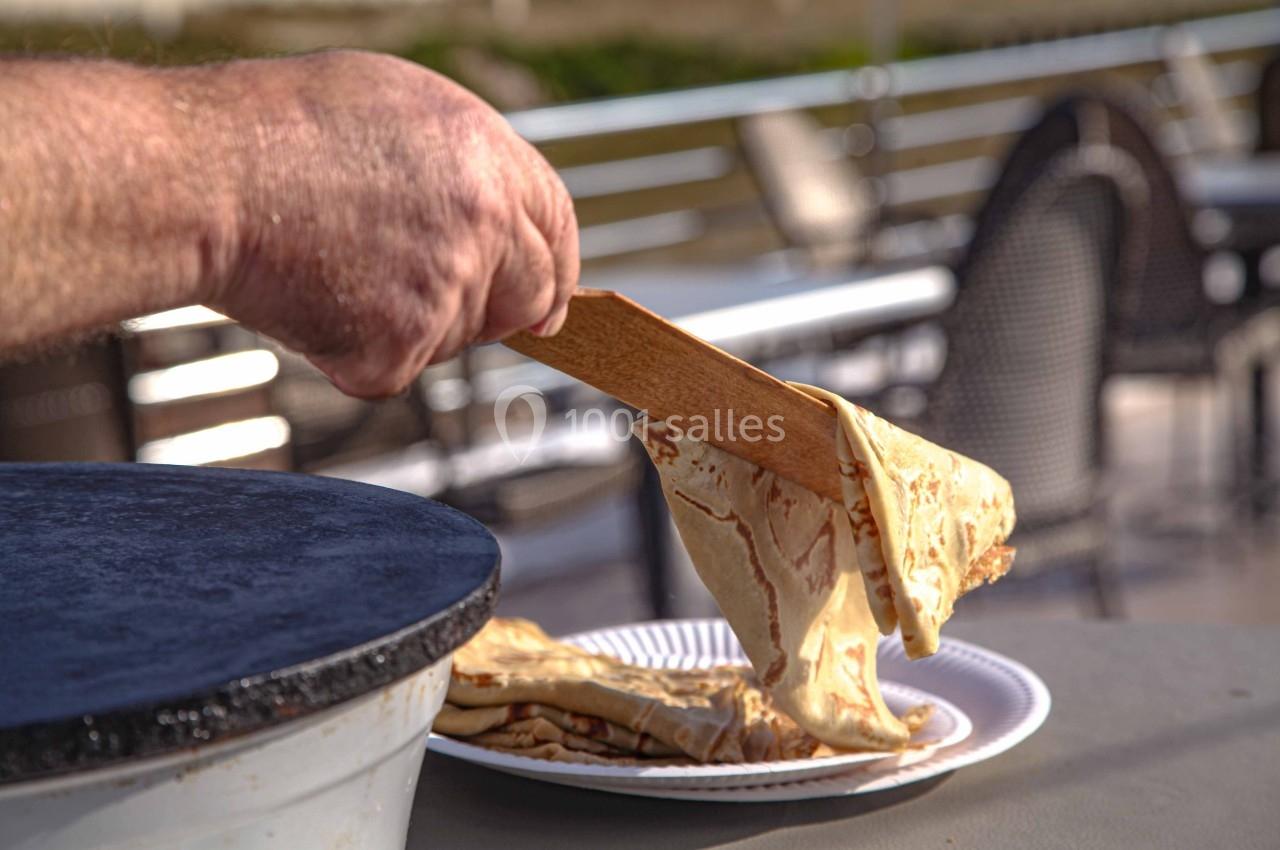 Une main dépose une crêpe chaude sur une assiette à l'aide d'une spatule, près d'une crêpière en extérieur.