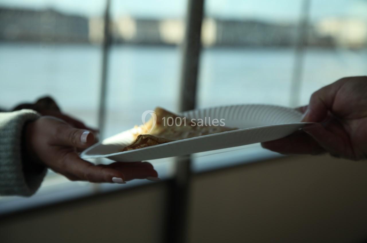 Une personne tend une assiette en carton contenant une crêpe à une autre personne, en arrière-plan flou d'une fenêtre.