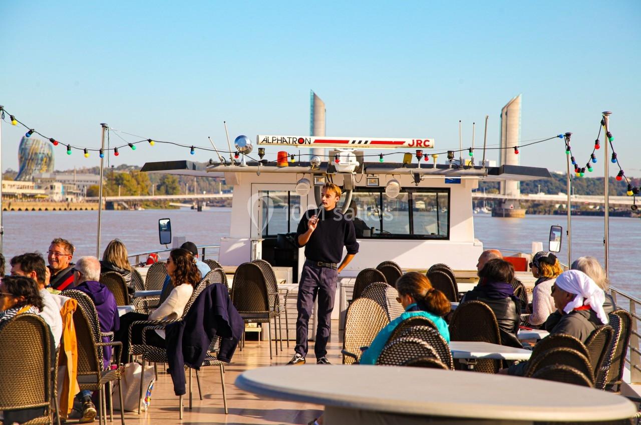 Un guide parle à un groupe de passagers assis sur le pont d'un bateau naviguant sur un fleuve par temps ensoleillé.