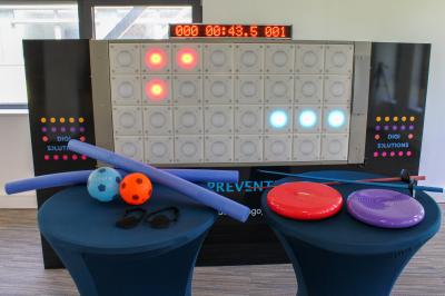 Miniature Multitudes de jeux sur le mur interactif  Mur interactif lumineux avec chronomètre, accessoires de sport et équilibre disposés sur deux tables bleues au premier plan.