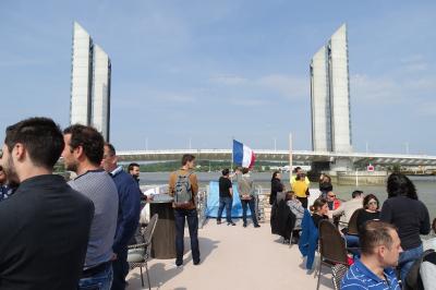 Miniature Burdigala II sous le Pont Jacques Chaban Delmas Bâtiment moderne en verre et métal près d'un fleuve, avec un bateau blanc amarré au premier plan.
