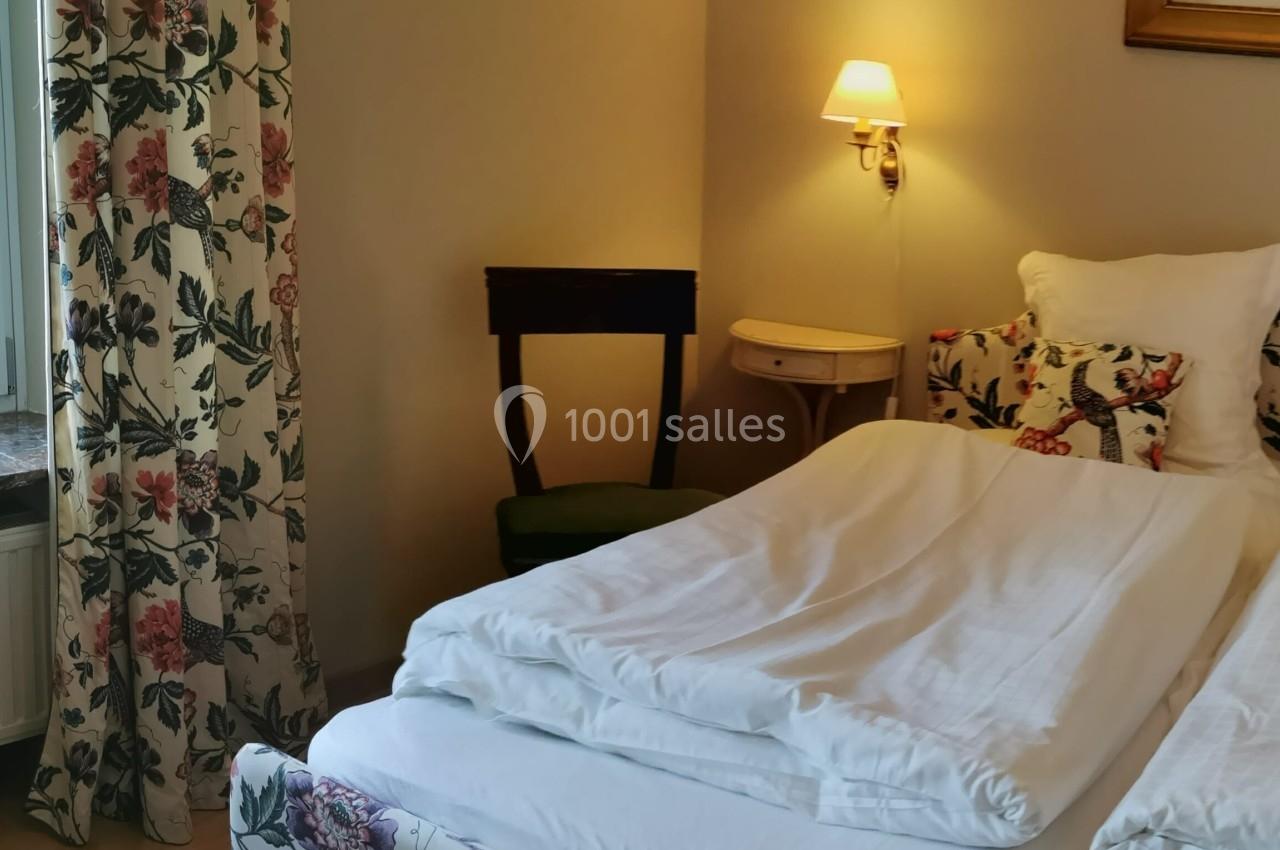 Lit double avec draps blancs dans une chambre décorée de motifs floraux, éclairée par une lampe murale.