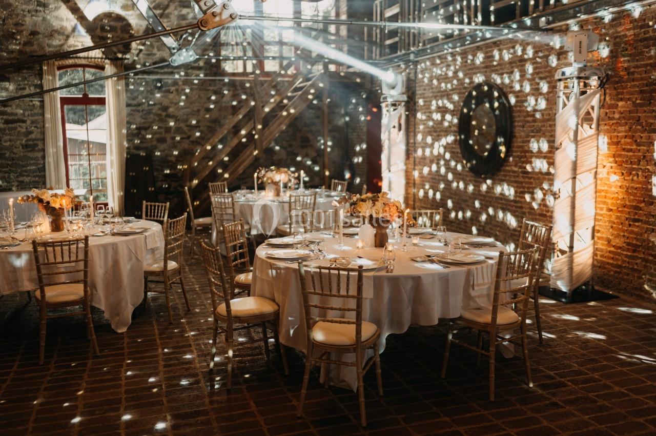 Salle de réception décorée avec des tables rondes, nappes blanches, chaises en bois clair et éclairage tamisé.