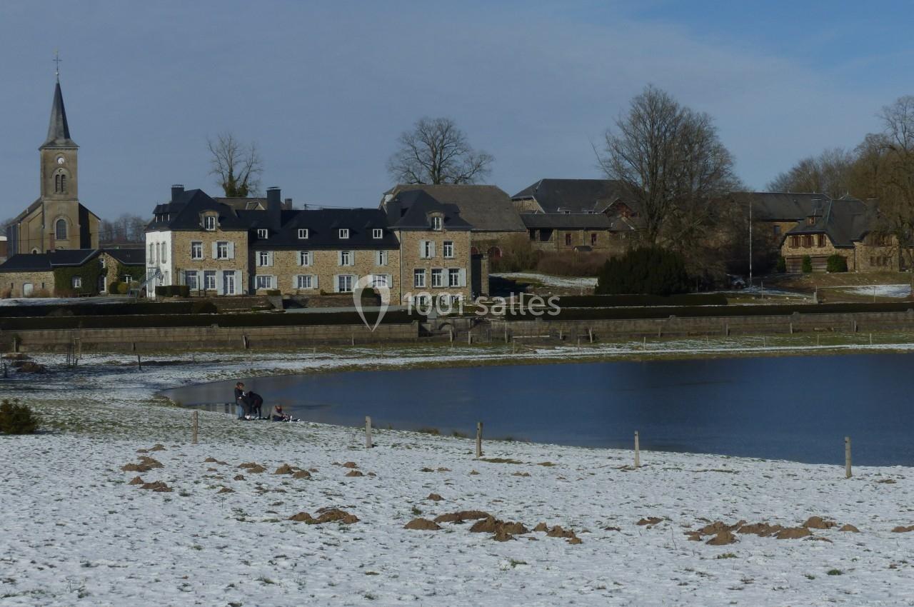 Village enneigé avec un étang gelé, des maisons en pierre, une église et des arbres dénudés sous un ciel bleu.