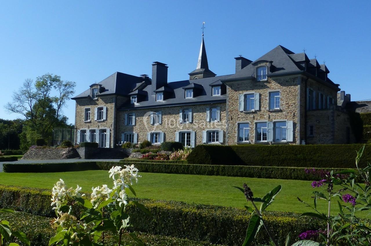 Manoir en pierre entouré de jardins bien entretenus, avec ciel bleu en arrière-plan.