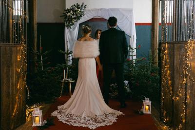 Miniature Célébration dans les box de la Salle Hermès Un couple en tenue de mariage se tient devant un officiant dans un décor festif avec sapins et guirlandes lumineuses.