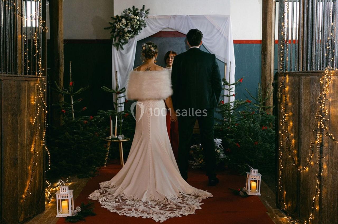Un couple en tenue de mariage se tient devant un officiant dans un décor festif avec sapins et guirlandes lumineuses.