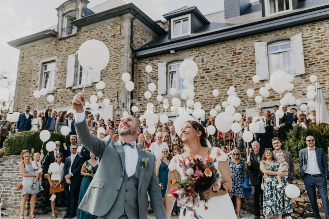 Un couple de mariés souriants devant une foule, libérant des ballons blancs devant une maison en pierre.
