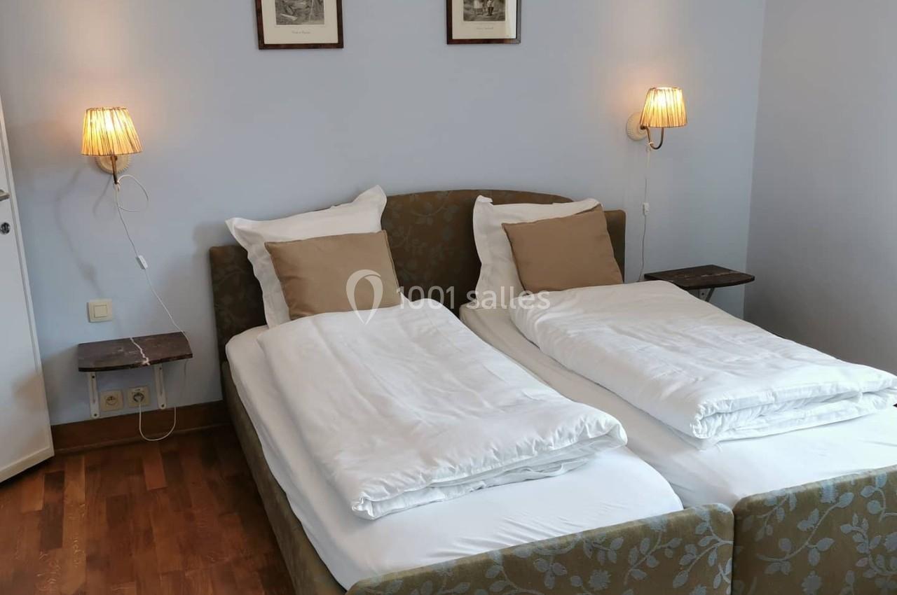 Deux lits simples avec linge blanc, tête de lit fleurie, lampes murales et parquet en bois dans une chambre sobre.