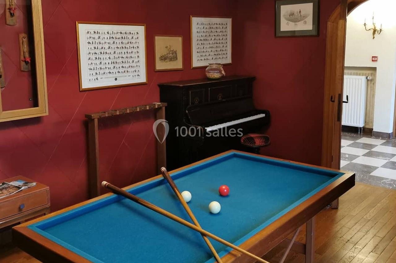 Table de billard français avec deux queues croisées, trois billes et un piano en arrière-plan dans une pièce décorée.