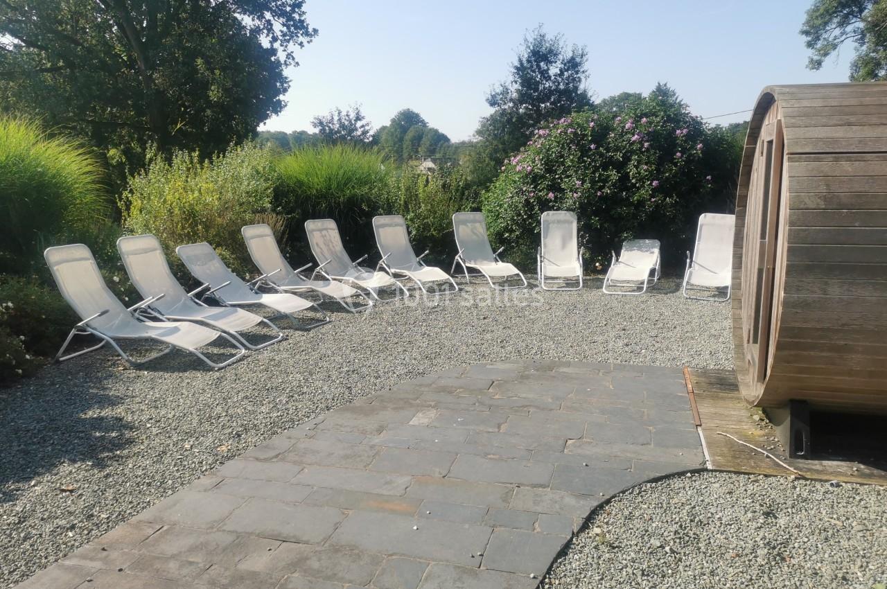 Chaises longues blanches disposées en arc de cercle sur du gravier, entourées de végétation, près d'un sauna en bois.