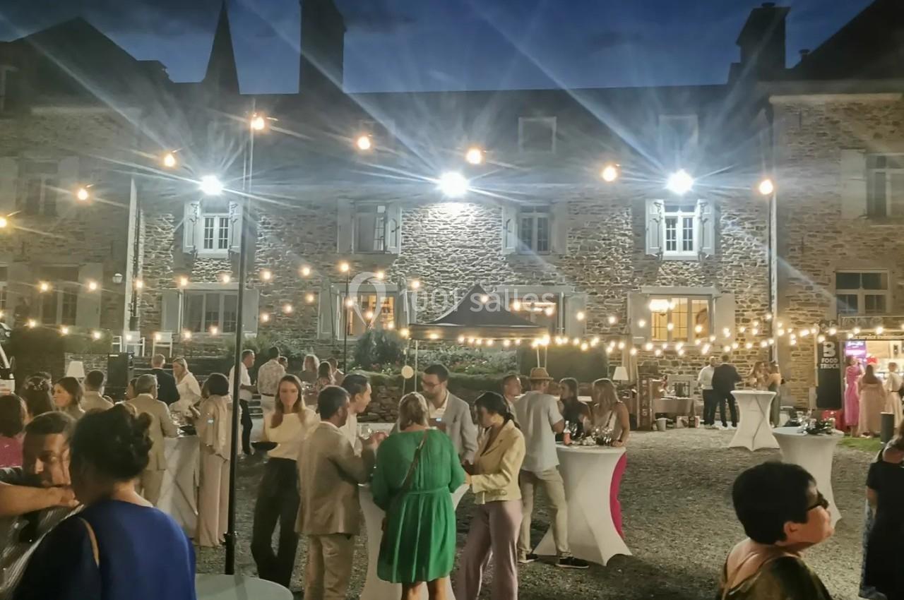 Groupe de personnes discutant lors d'une réception en plein air, éclairée par des guirlandes lumineuses devant un bâtiment…