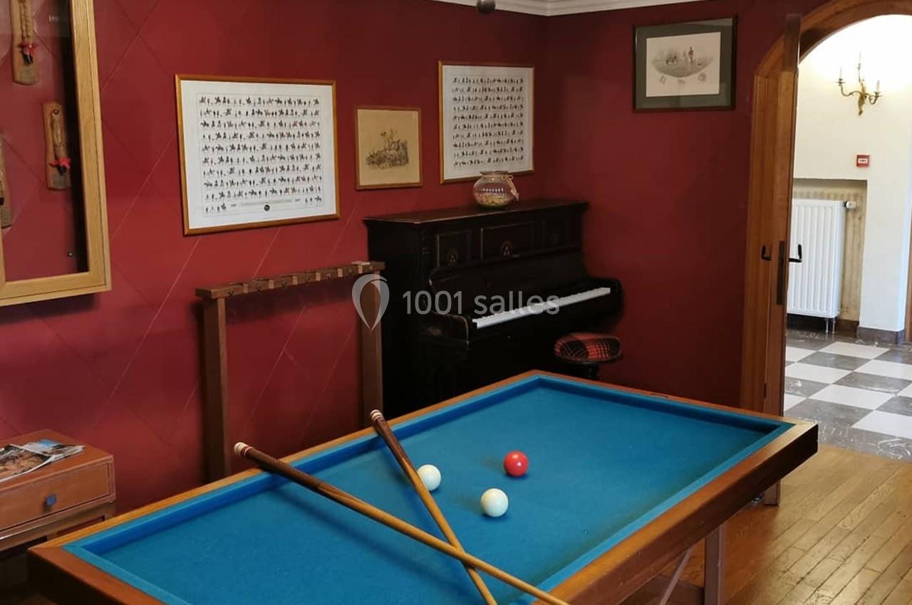 Table de billard avec trois boules et deux queues, dans une pièce avec un piano, des cadres et des murs rouges.