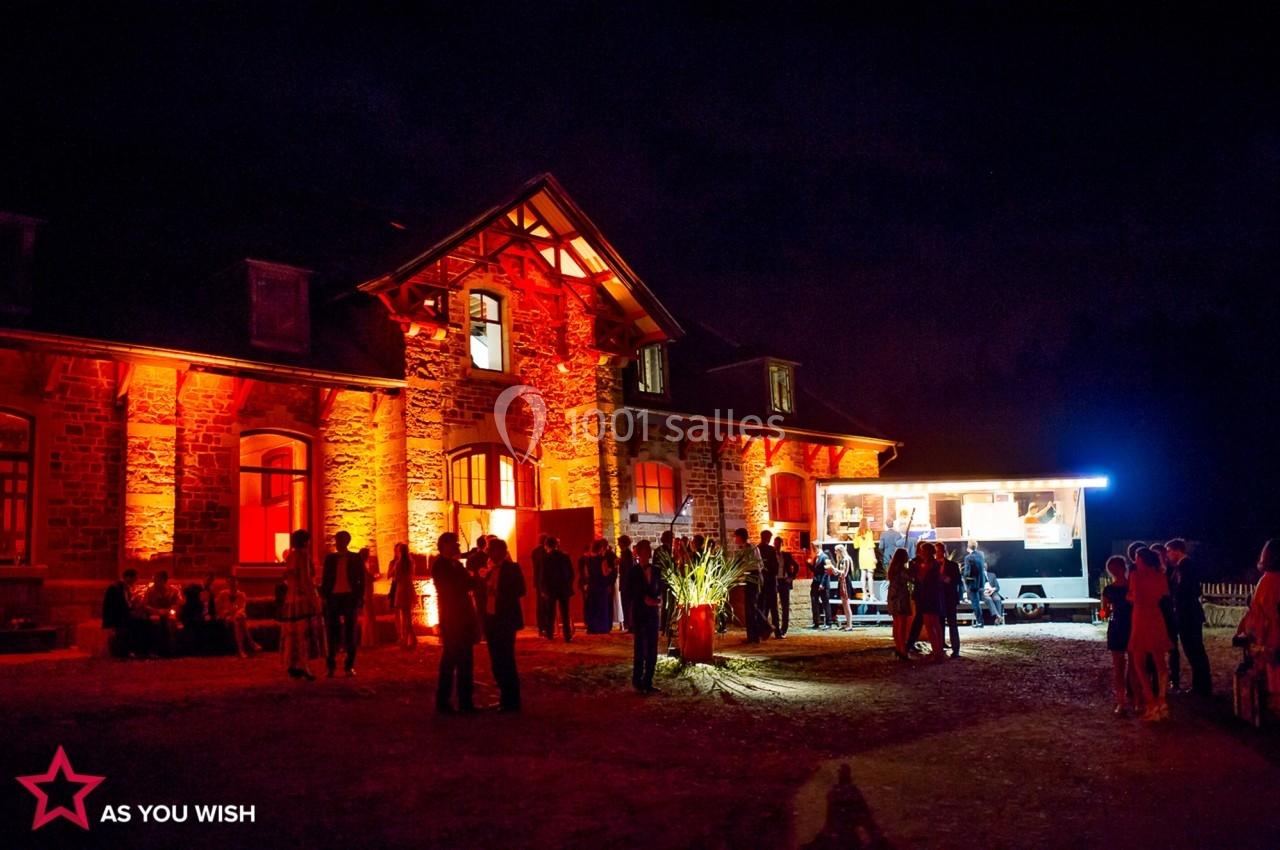 Façade d'un bâtiment en pierre éclairée en rouge, avec un food truck et des groupes de personnes rassemblées la nuit.