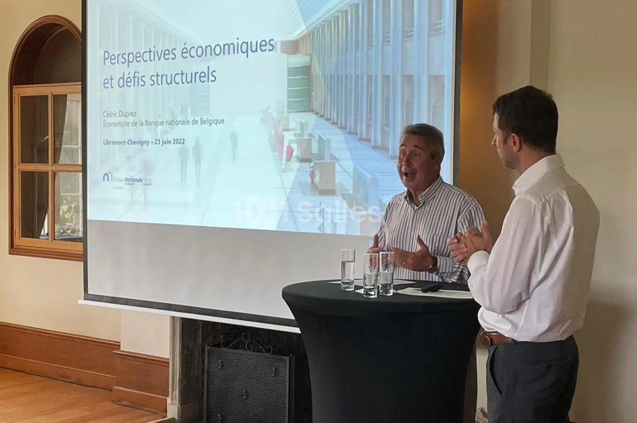 Deux hommes discutent devant un écran projetant une présentation sur les perspectives économiques dans une salle lumineuse.