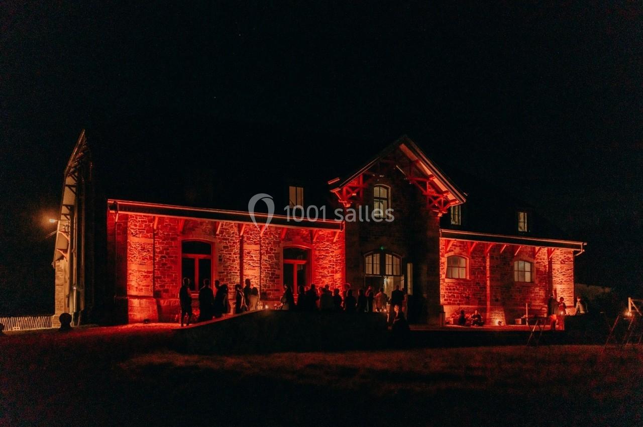 Façade d'une grande maison en pierre éclairée par des lumières rouges, avec des silhouettes de personnes devant.