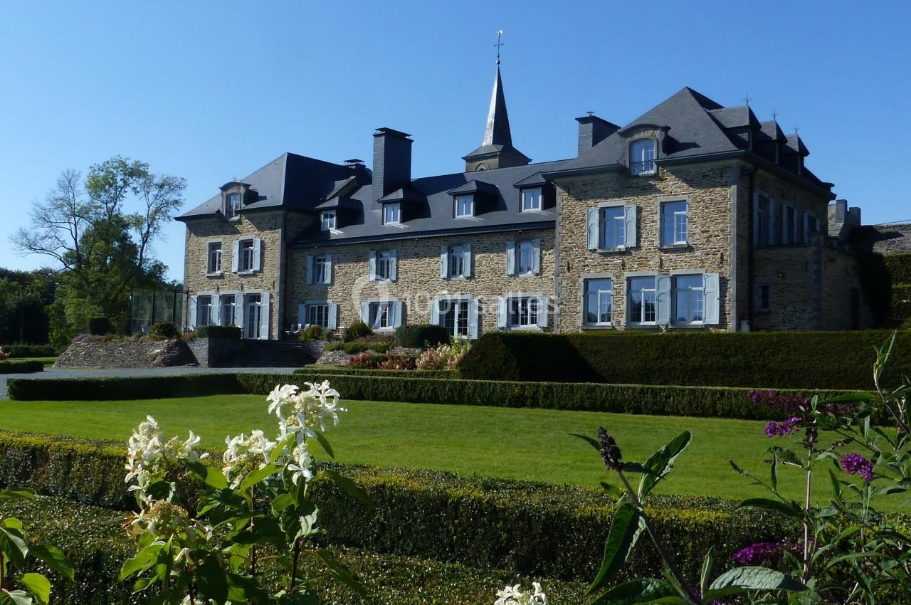 Manoir en pierre entouré de jardins paysagers avec haies taillées, fleurs et pelouse verdoyante.