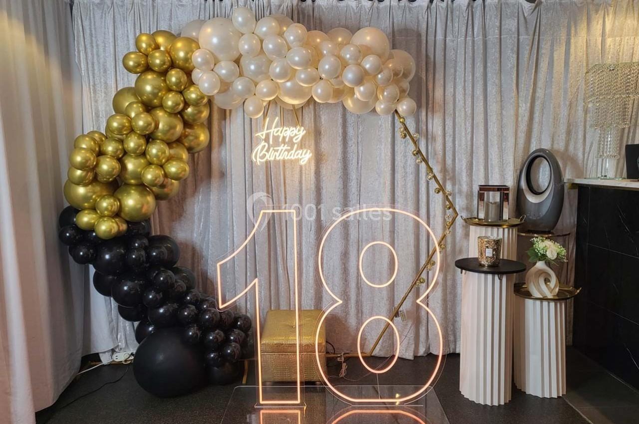 Décoration d'anniversaire avec ballons dorés, noirs et blancs, grand chiffre ’18’ lumineux et rideau argenté en fond.