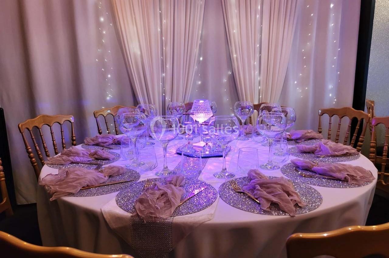 Table ronde élégamment dressée avec nappes blanches, serviettes pliées, verres et décorations lumineuses violettes.