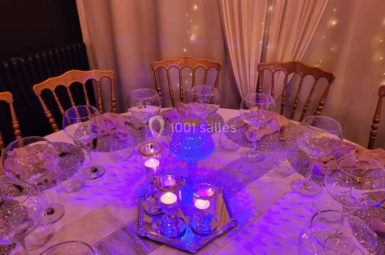 Table élégamment dressée avec nappes blanches, verres, bougies et éclairage violet dans une ambiance festive.