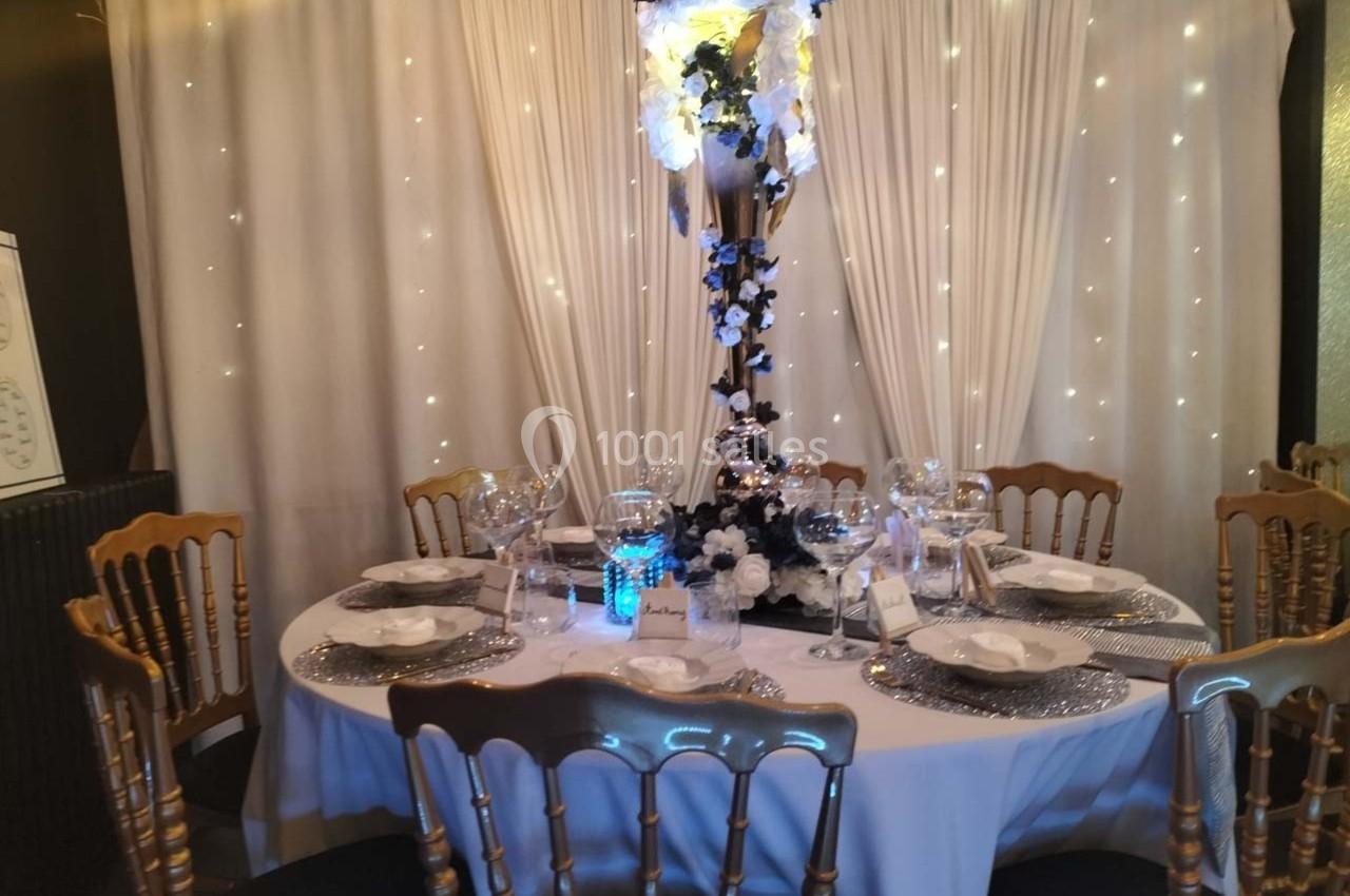 Table ronde élégamment dressée avec nappes blanches, chaises dorées, centre de table floral et rideau lumineux en arrière…