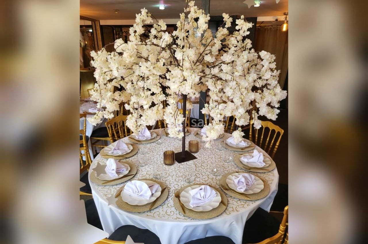 Table ronde décorée avec une nappe blanche, des assiettes dorées, des serviettes pliées et un centre floral blanc élégant.