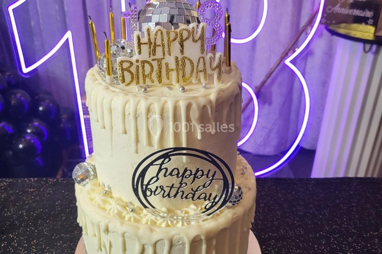 Gâteau d'anniversaire à deux étages décoré de glaçage blanc, bougies dorées et ornements ’Happy Birthday’.