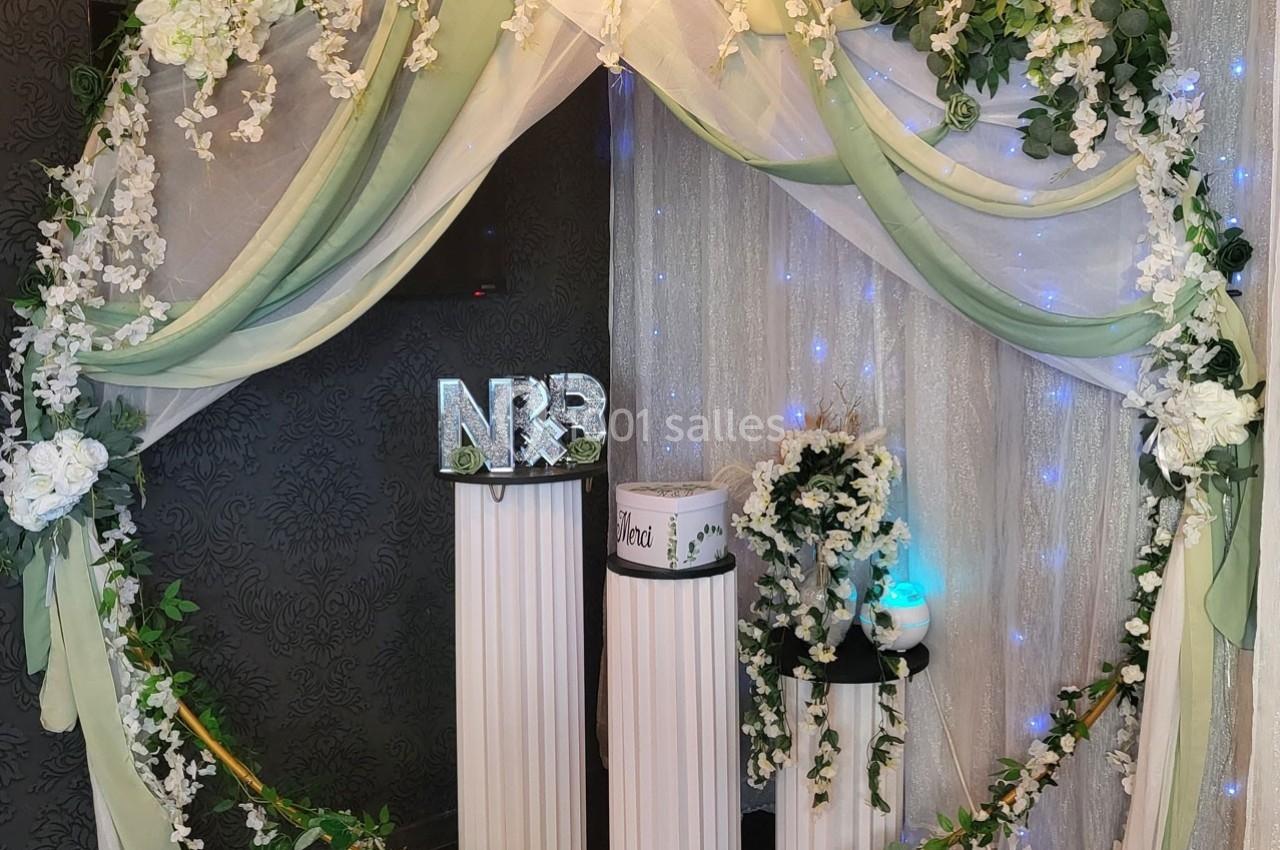 Décoration de cérémonie avec arche florale, rideaux lumineux, colonnes blanches et initiales ’NR’ en avant-plan.