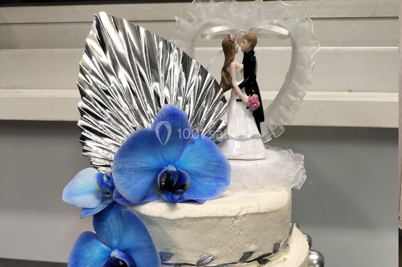 Figurines de mariés sur un gâteau décoré de fleurs bleues, d'une feuille argentée et d'un cœur en arrière-plan.