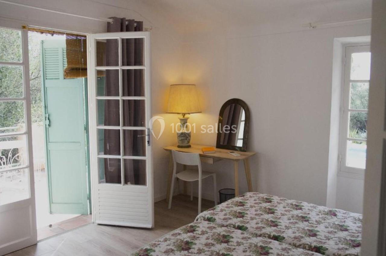 Chambre lumineuse avec lit à motifs floraux, bureau avec miroir, lampe allumée et porte-fenêtre ouverte sur une terrasse.