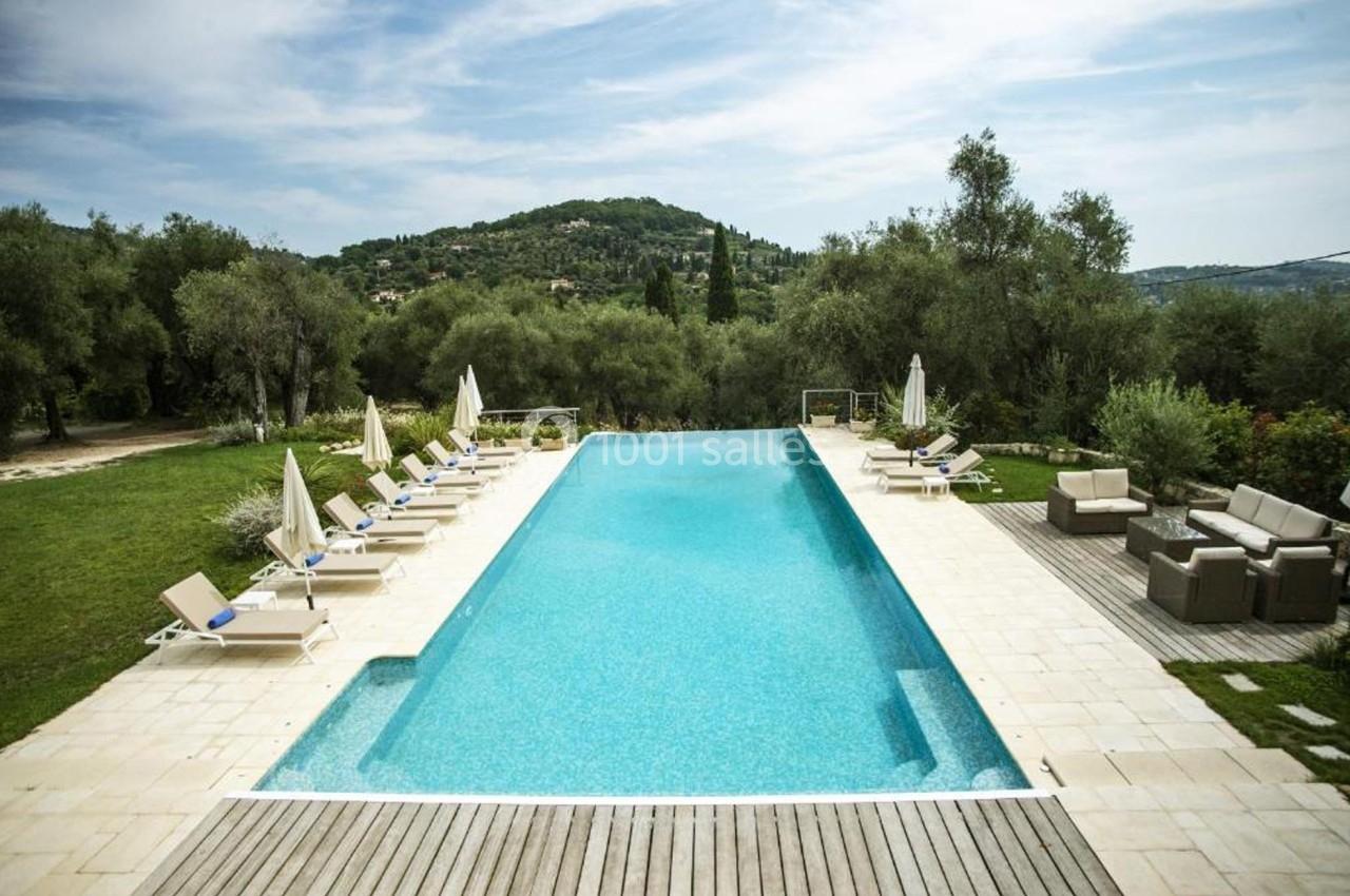 Piscine extérieure entourée de transats et parasols, avec vue sur un paysage verdoyant et colline en arrière-plan.