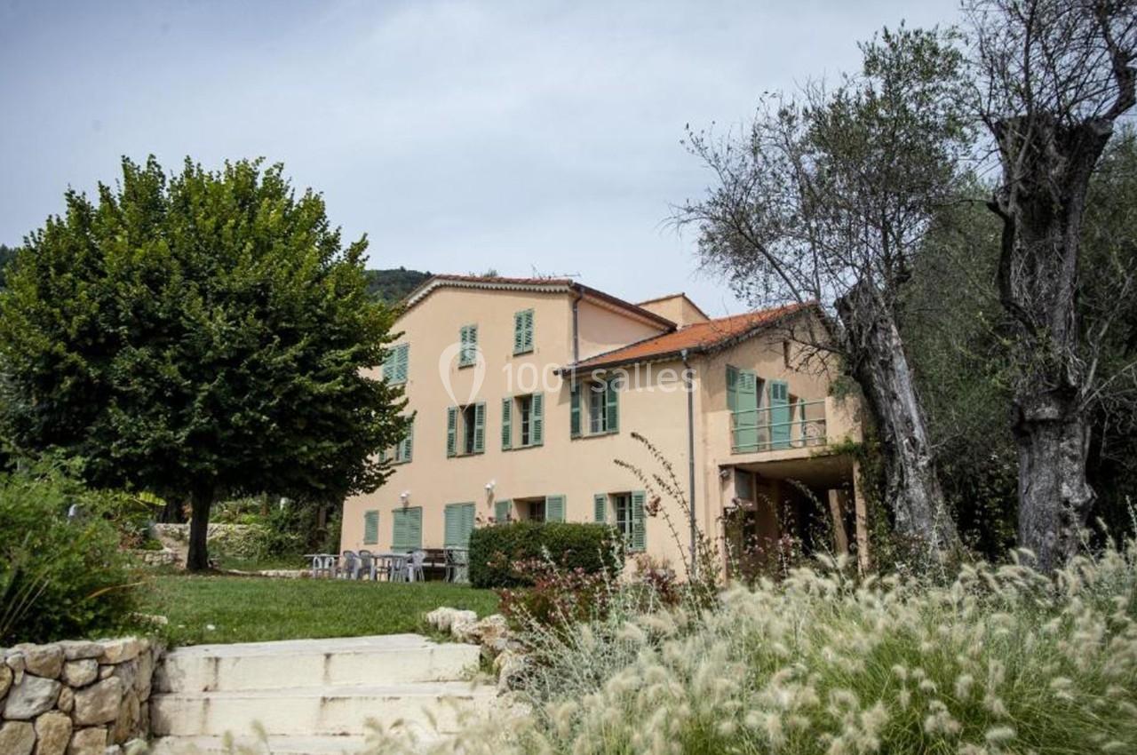 Grande maison beige à volets verts entourée de végétation, avec un escalier en pierre menant à un jardin.