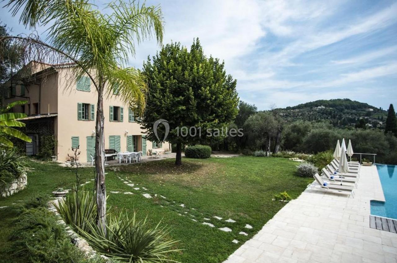 Maison beige avec volets verts, jardin arboré, piscine à débordement et vue sur une colline verdoyante.