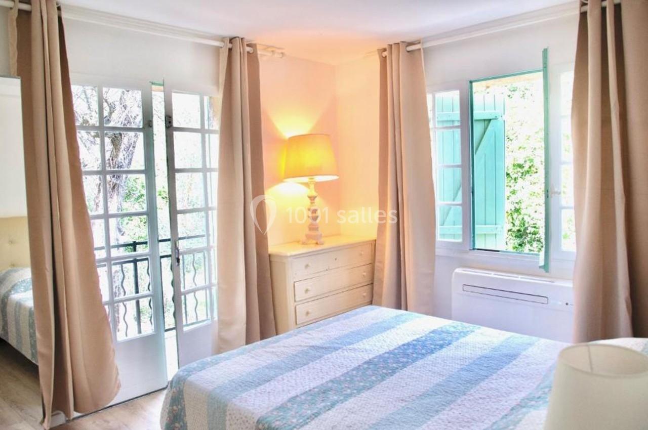 Chambre lumineuse avec lit double, lampadaire allumé, rideaux beige, fenêtres ouvertes sur un jardin verdoyant.