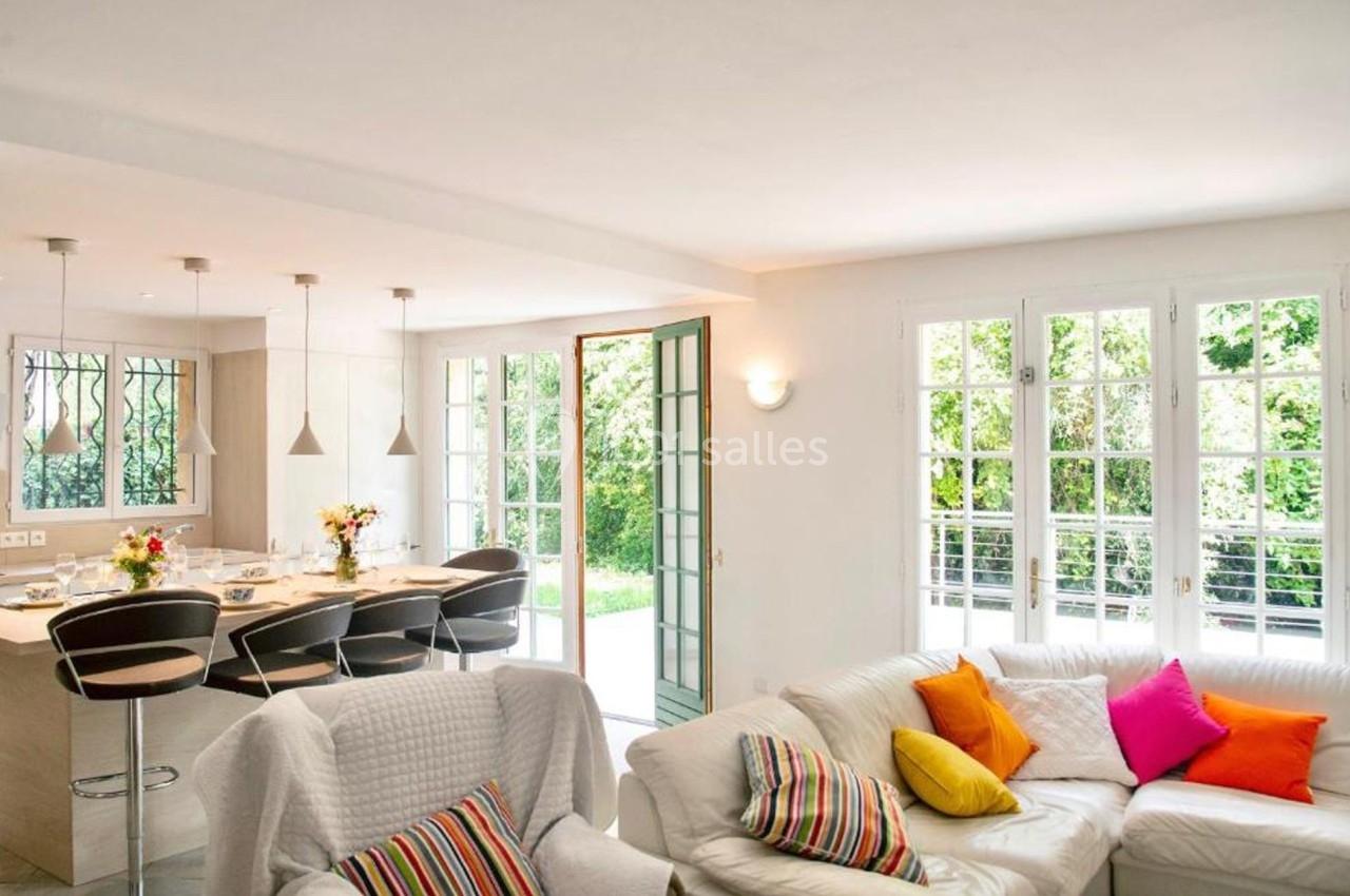 Salon lumineux avec canapé blanc et coussins colorés, ouvert sur une cuisine moderne et un jardin visible par de grandes…