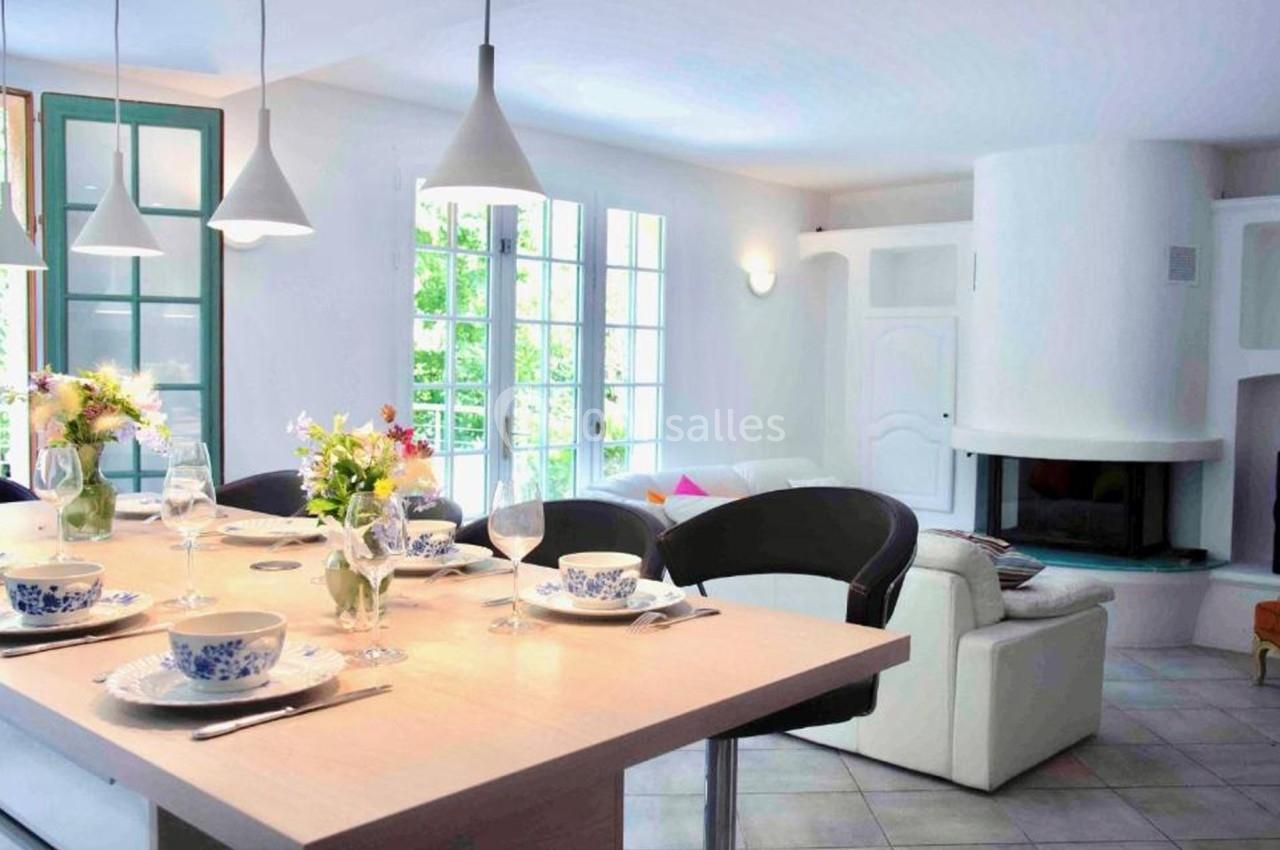 Salle à manger lumineuse avec table dressée, chaises noires, cheminée blanche et grandes fenêtres donnant sur un jardin.
