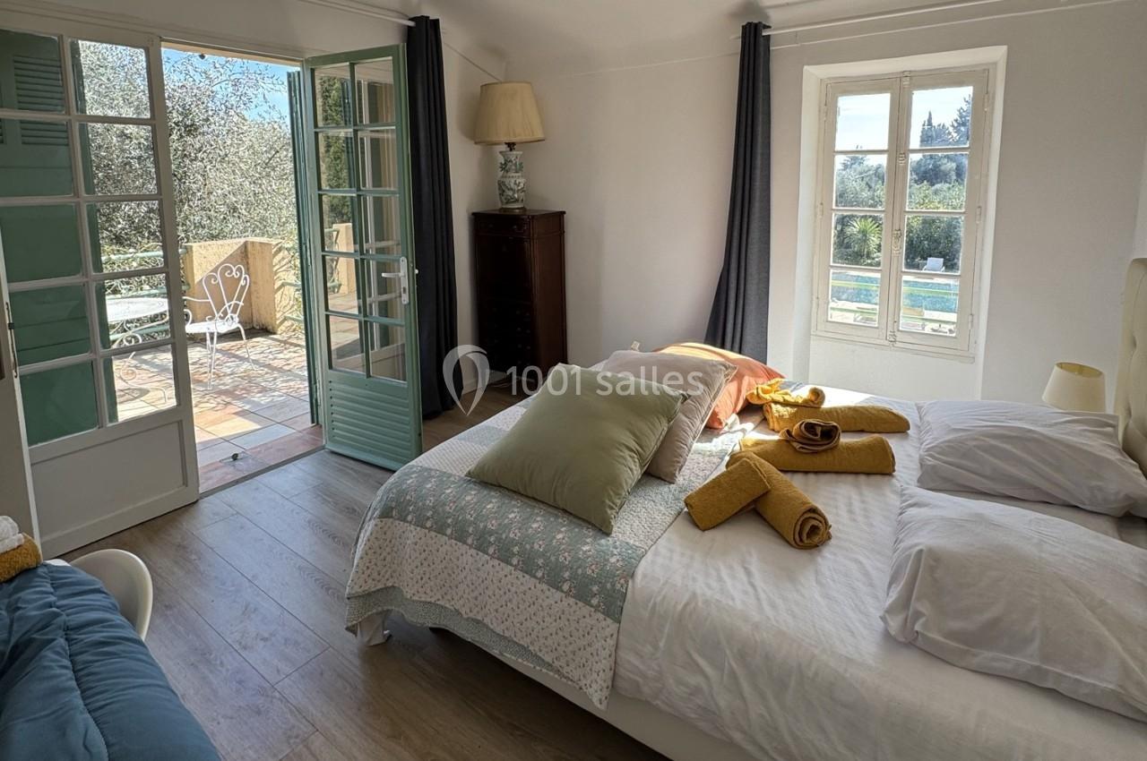 Chambre lumineuse avec lit double, draps et serviettes jaunes, donnant sur une terrasse ensoleillée avec vue sur un jardin.