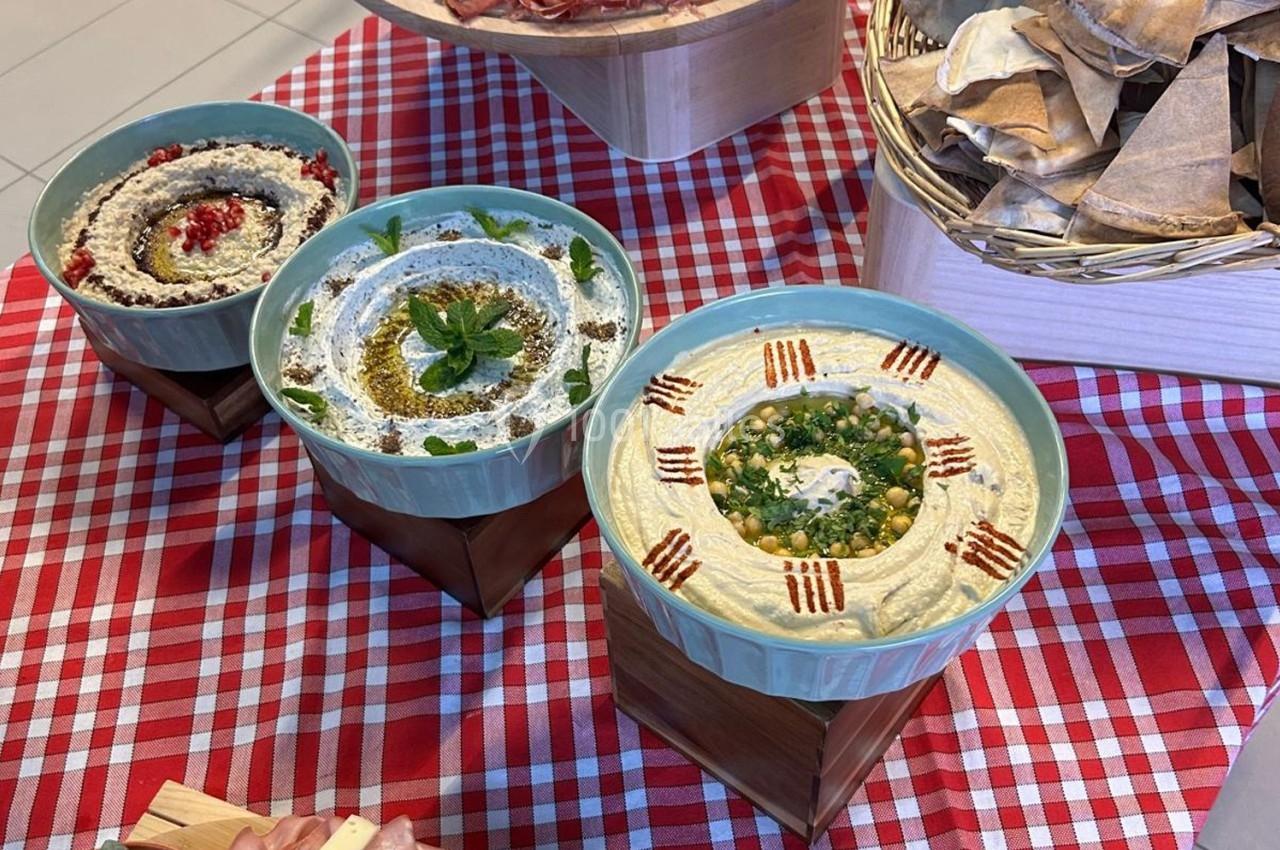 Trois bols de houmous garnis d'herbes, épices et pois chiches, présentés sur une nappe à carreaux rouges et blancs.
