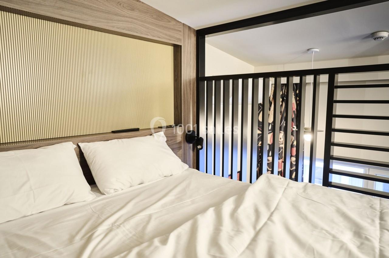 Lit double avec draps blancs dans une chambre moderne, séparée par une cloison en bois et une rambarde métallique.