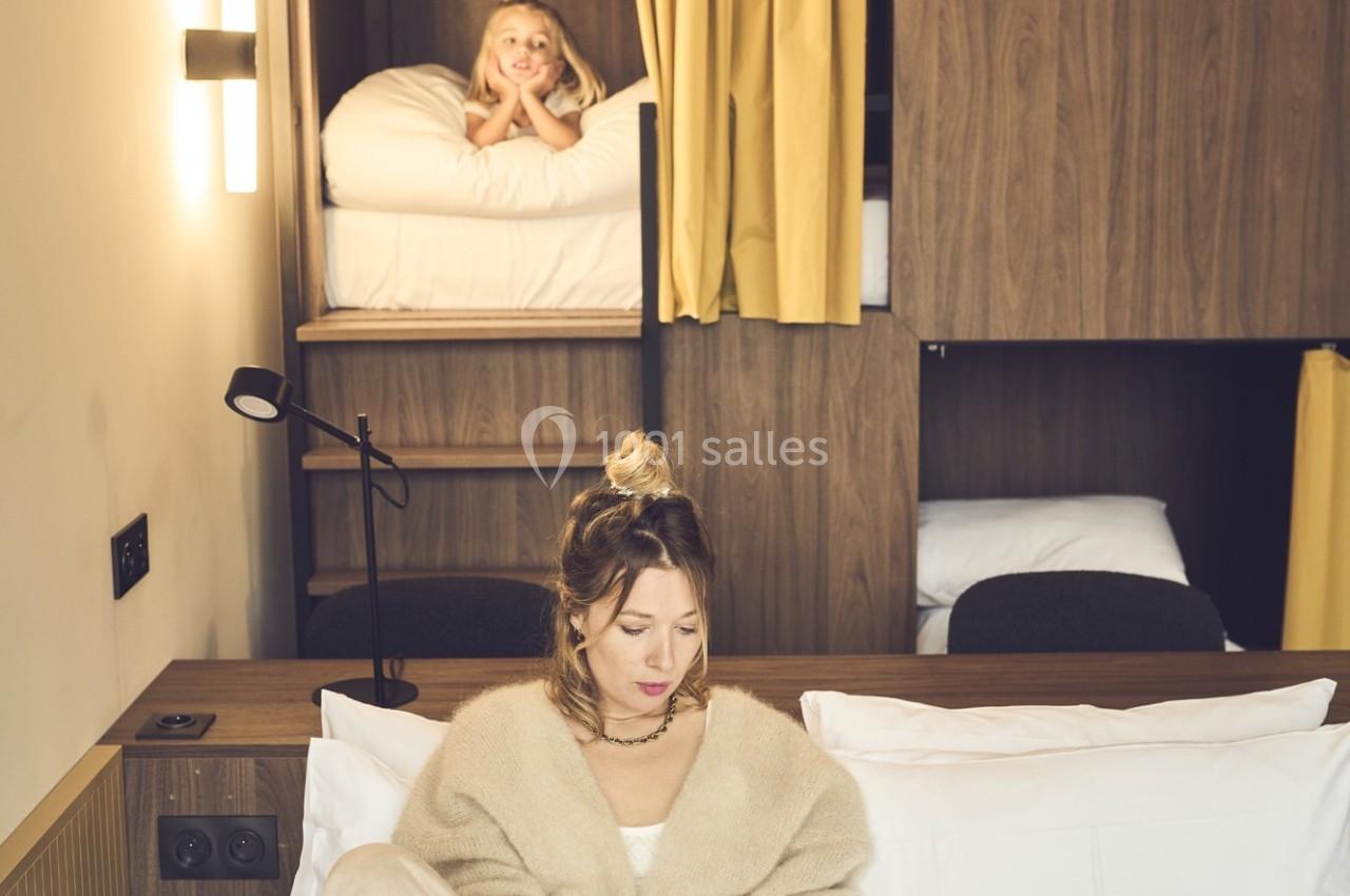 Une femme assise sur un lit lit un document, tandis qu'une enfant sourit depuis une couchette en arrière-plan.