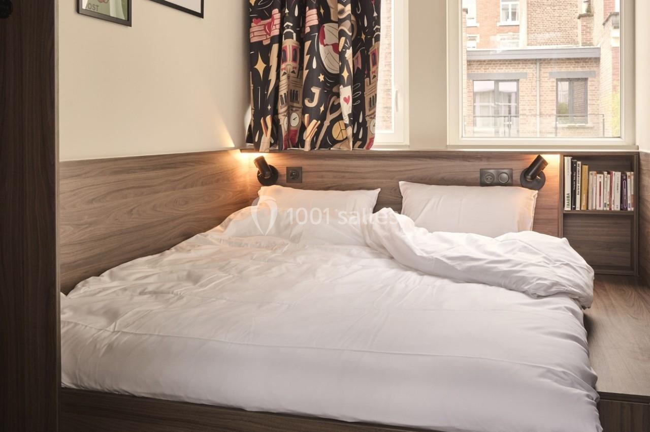 Lit double avec draps blancs dans une chambre lumineuse, décorée de rideaux colorés et d'étagères avec des livres.