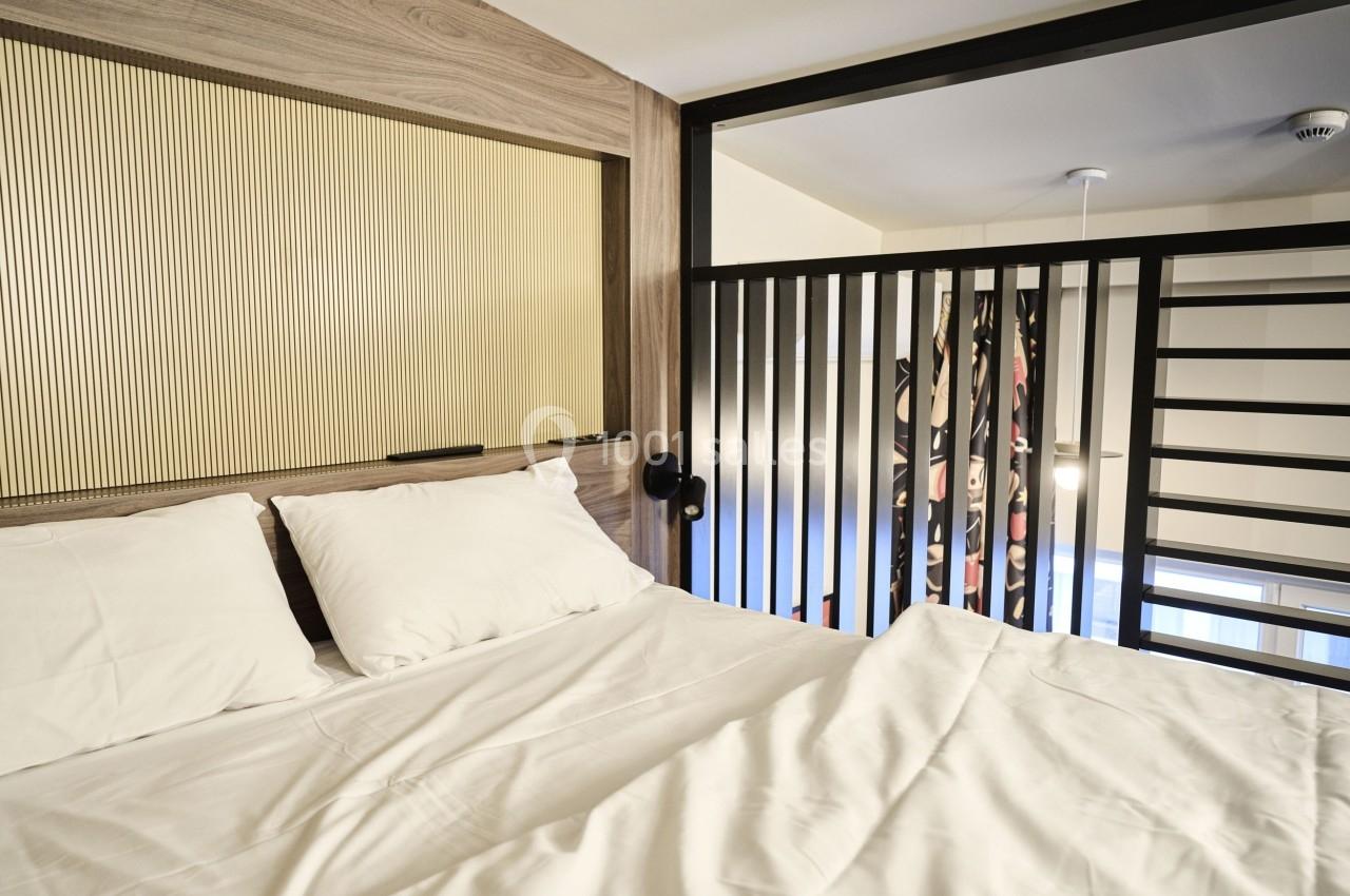 Lit double avec draps blancs dans une chambre moderne, séparée par une cloison en bois et une rambarde métallique.