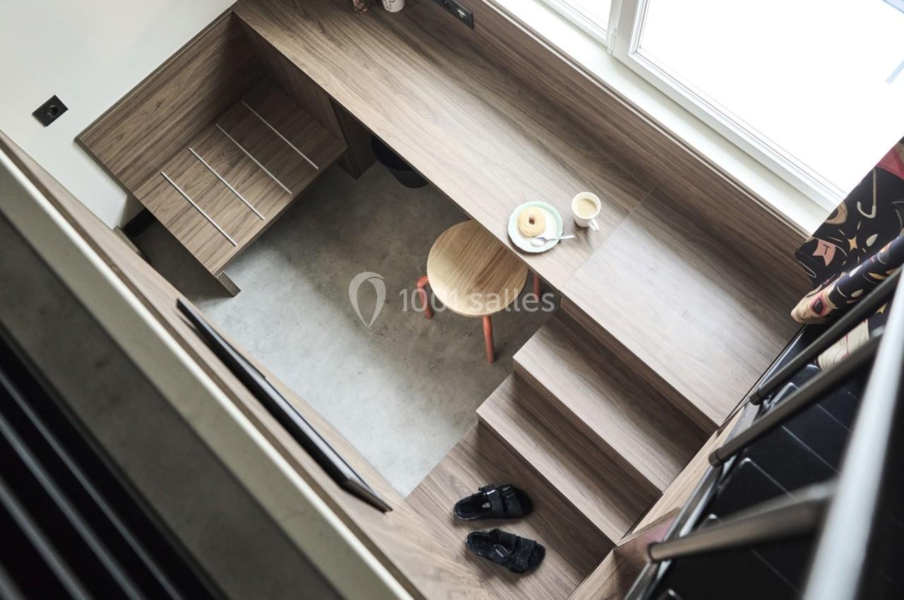 Vue plongeante sur un espace de travail compact avec bureau en bois, tabouret, tasse, assiette et escaliers intégrés.