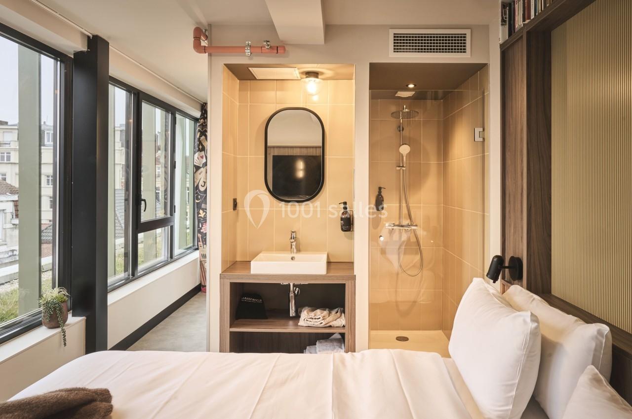 Chambre lumineuse avec lit, salle de bain ouverte comprenant une douche, un lavabo et un miroir, vue sur des fenêtres.