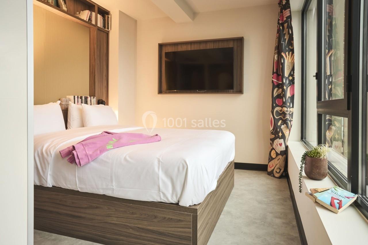 Chambre lumineuse avec lit double, couverture rose, étagères intégrées, télévision murale et grande fenêtre avec rideaux…